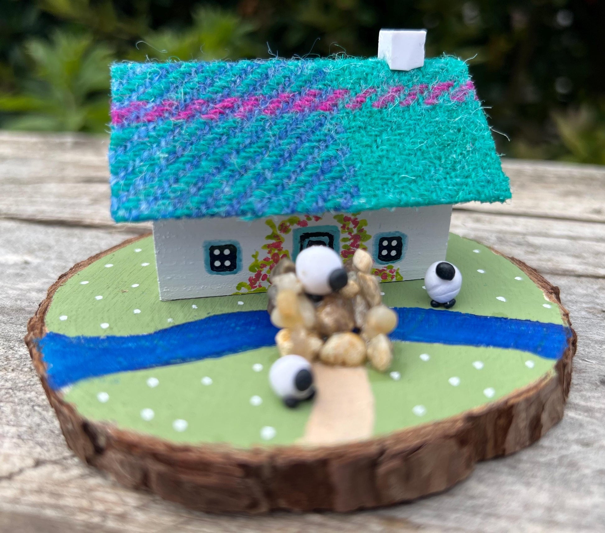 white handpainted wooden cottage with a teal and pink harris tweed roof.  The cottage has a light blue door and windows and there are red flowers painted around the door.  The cottage is on a green painted base with a painted river and a stone bridge.  There are three miniature sheep on the grass outside the cottage. 