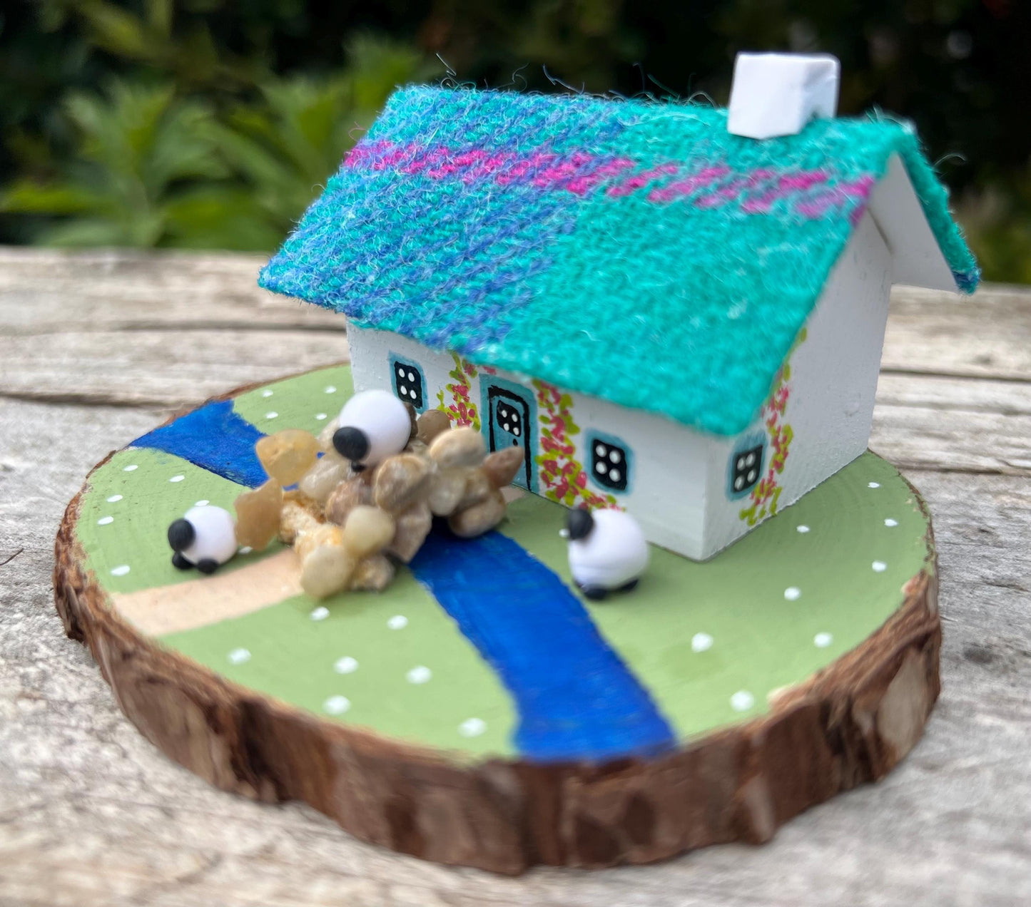 white handpainted wooden cottage with a teal and pink harris tweed roof.  The cottage has a light blue door and windows and there are red flowers painted around the door.  The cottage is on a green painted base with a painted river and a stone bridge.  There are three miniature sheep on the grass outside the cottage. 