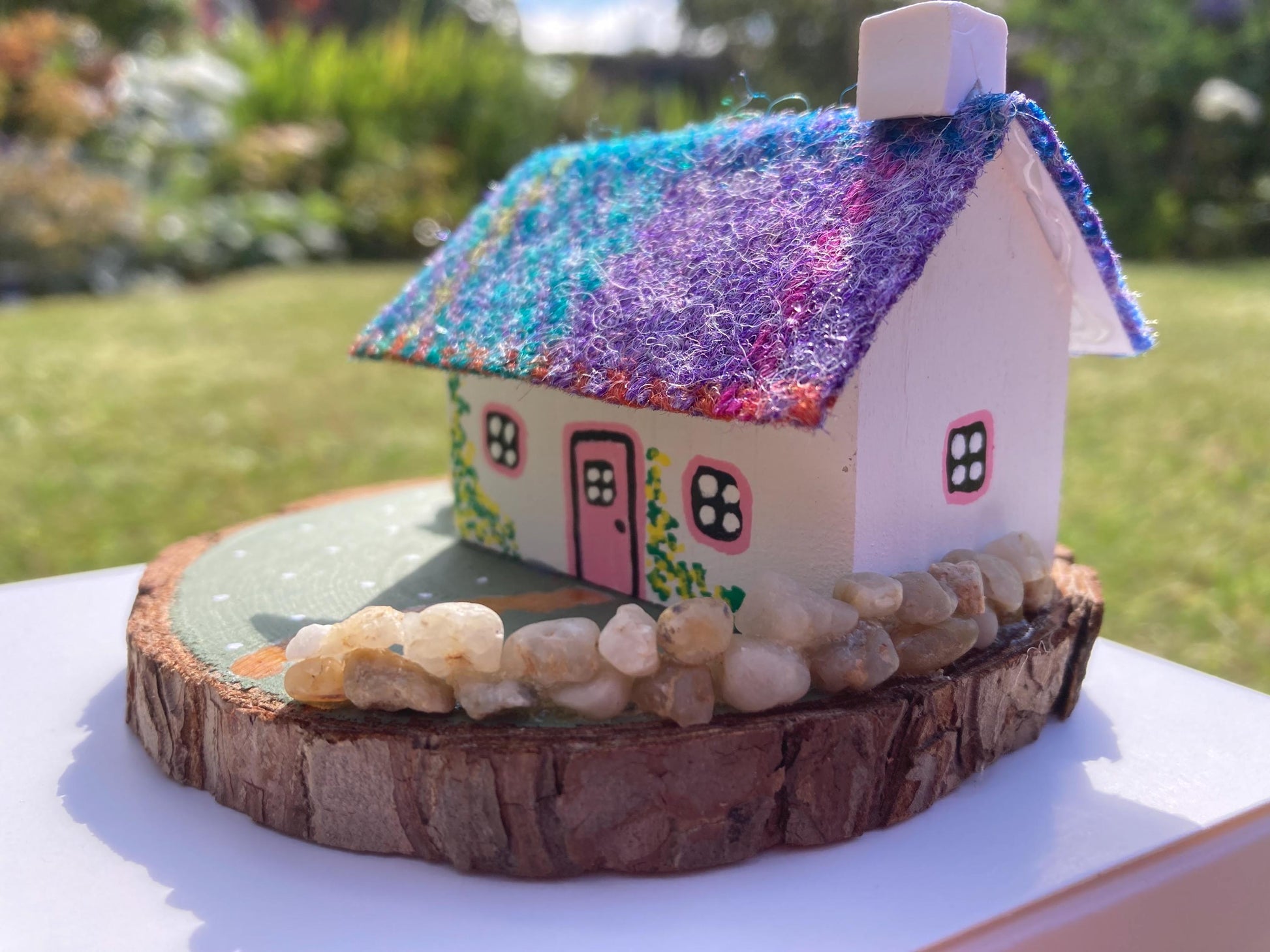 Miniature model of a white scottish bothy with stone built log store on a green painted base featuring a path and stone wall.   The  Cottage has a pink door and windows with yellow flowers on the walls around the door. There is purple and teal harris tweed on the roof.  