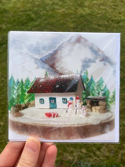 A printed square greetings card feauturing a snow covered highland bothy with a red harris tweed roof, teal doors and windows.  There are pine trees and mountains in the background.  Outside there are three snowmen of different sizes and a red sled.  There is a stone log store at the side of the cottage. 