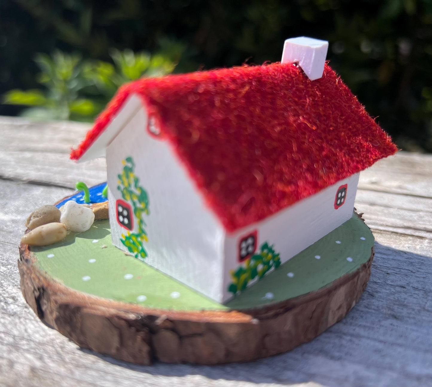 White wooden handpainted cottage with red harris tweed roof.  The cottage has a red door and windows and there are yellow flowers painted around the door and on the walls.  The cottage sits on a green base with a painted lake.  There is a model loch ness monster swimming in the lake. 