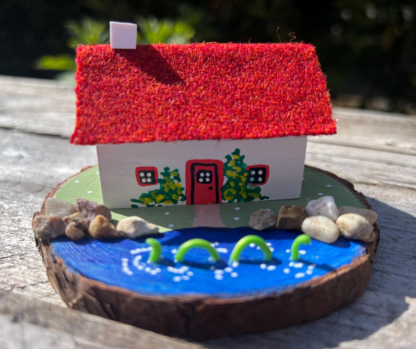 White wooden handpainted cottage with red harris tweed roof.  The cottage has a red door and windows and there are yellow flowers painted around the door and on the walls.  The cottage sits on a green base with a painted lake.  There is a model loch ness monster swimming in the lake. 
