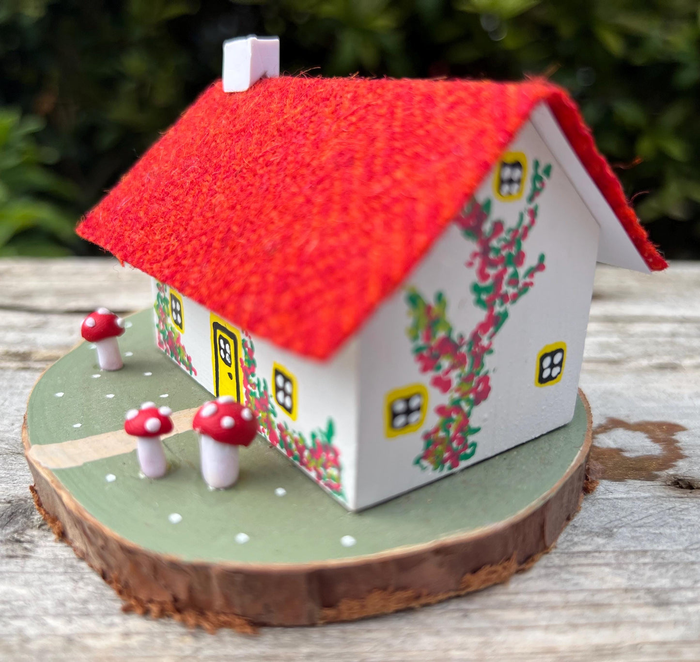 White hand painted cottage with red Harris tweed roof and red and white fairy mushrooms on the grass outside.  The cottage has yellow doors and windows and red flowers painted around the door.